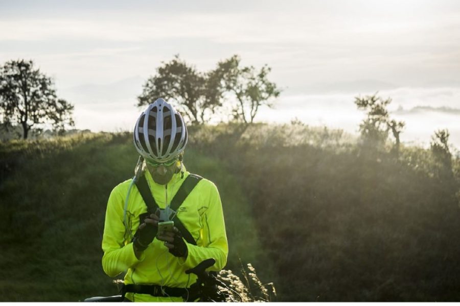 A bicycle rider wearing a bright yellow long-sleeved jacket and a helmet looks at their phone while on an outdoor trails lined with trees and tall grasses