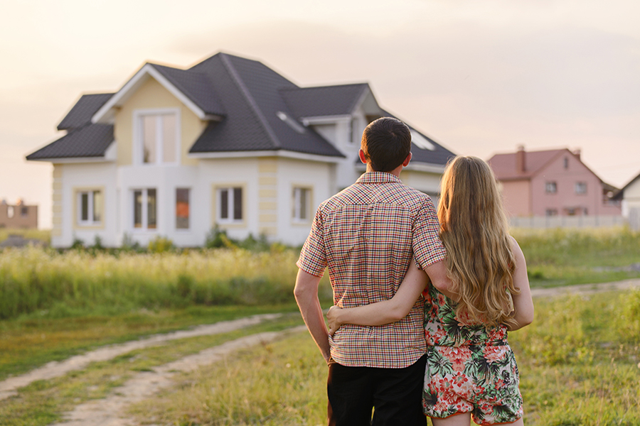 A young couple stands with their arms around each other, looking at a large single-family house in a grassy field