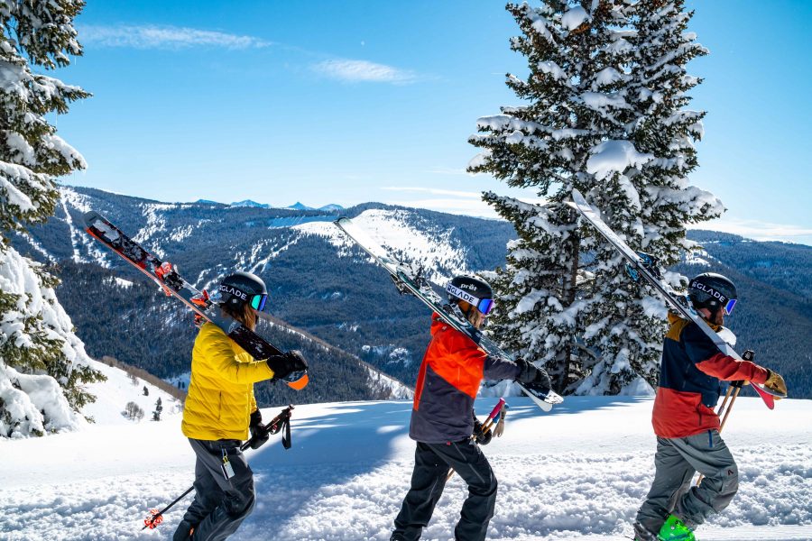 Three people wearing bright winter-weather clothing and carrying skis and poles walk in single-file along a snowy trail with snow covered mountains in the background