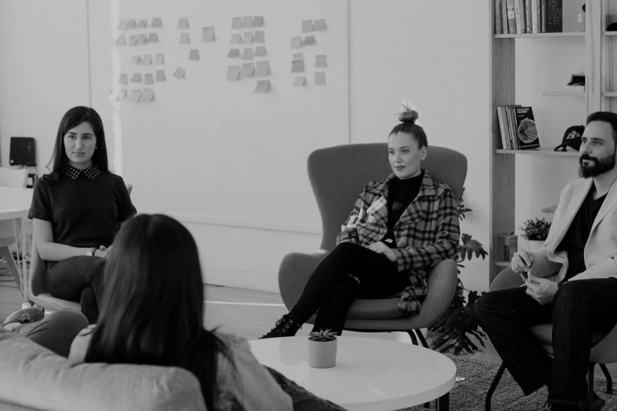 Four young professionals sit in a circle in an informal business meeting space with a white board in the background