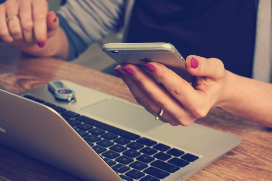 A woman sits at a desk with an open laptop in front of her and a smartphone in her hand