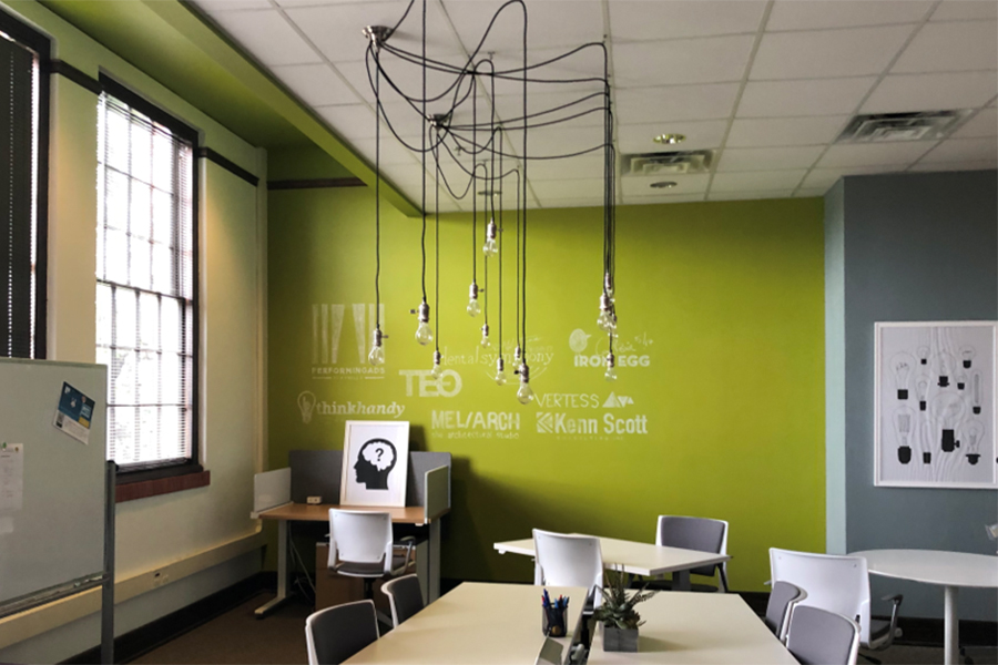 The inside of a small business office space with several desks, a conference table, a whiteboard, and bright yellow-green walls with inspirational words stenciled on it.