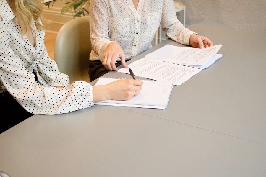 Two professional women sit next to each other at a table. One woman writes on a notepad while the other flips through a paper document