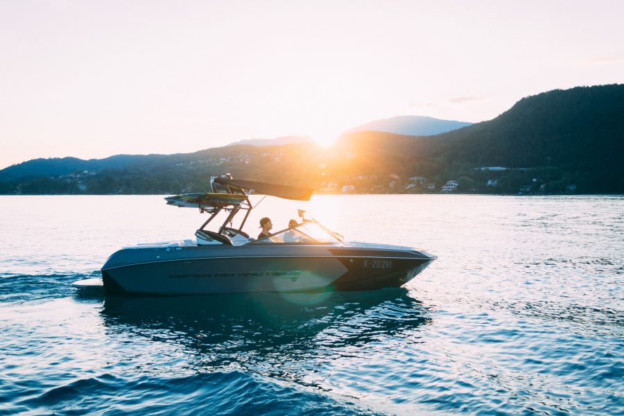 Two people cruise across a bay or large lake in a medium-sized motor boat at sunset. Hills dotted by houses can be seen in the distance.