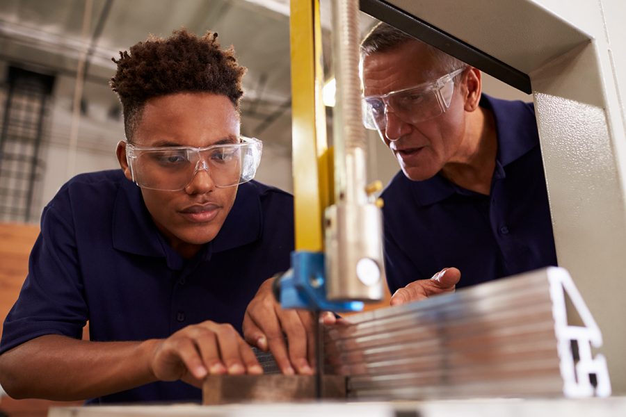 A young, black man wearing safety goggles uses a piece of equipment to cut pieces of metal while an older, white man wearing safety goggles provides instructions as part of a paid apprenticeship
