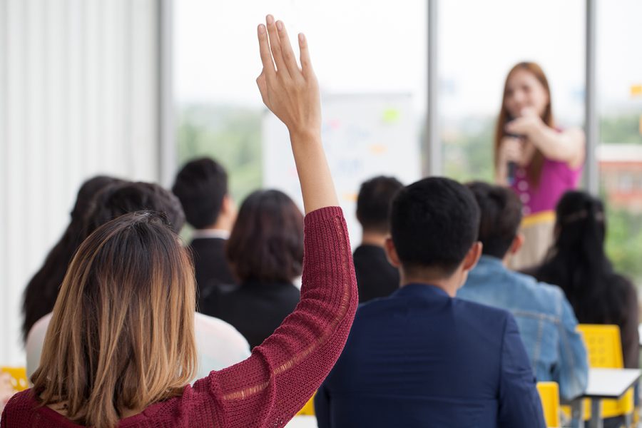 A group of people in a meeting room with moderator at the front of the room pointing to a person who has their hand raised