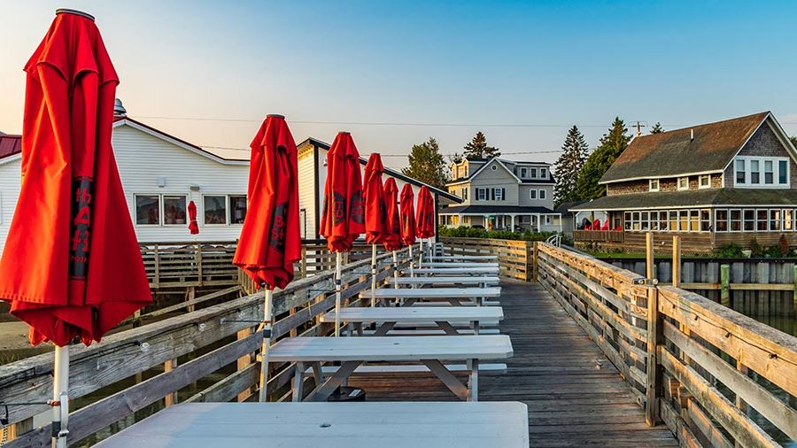 Outdoor seating area with a long row of picnic tables with bright reddish-orange umbrellas on a wooden waterfront dock in Scarborough, Maine.