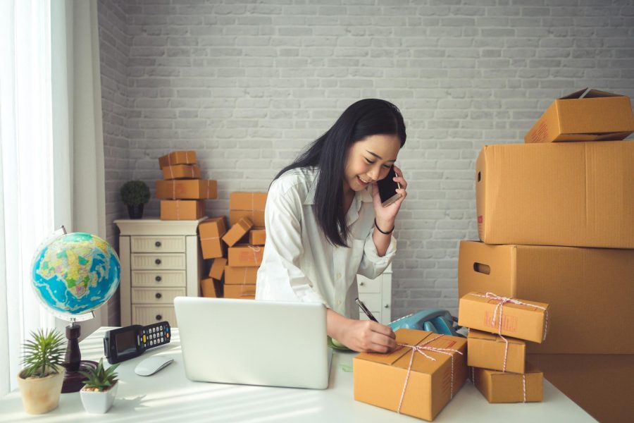 A young woman stands at a desk in her home office with her smartphone up to her ear. She has stacks of packages on her desk that she is writing addresses on. Additional stacks of boxes are behind her.