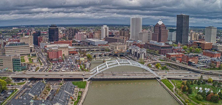 An aerial view of Rochester, New York, shows the Genesee River, the Frederick Douglass Susan B. Anthony Bridge, Court Street Dam, and many skyscrapers and smaller commercial and residential buildings