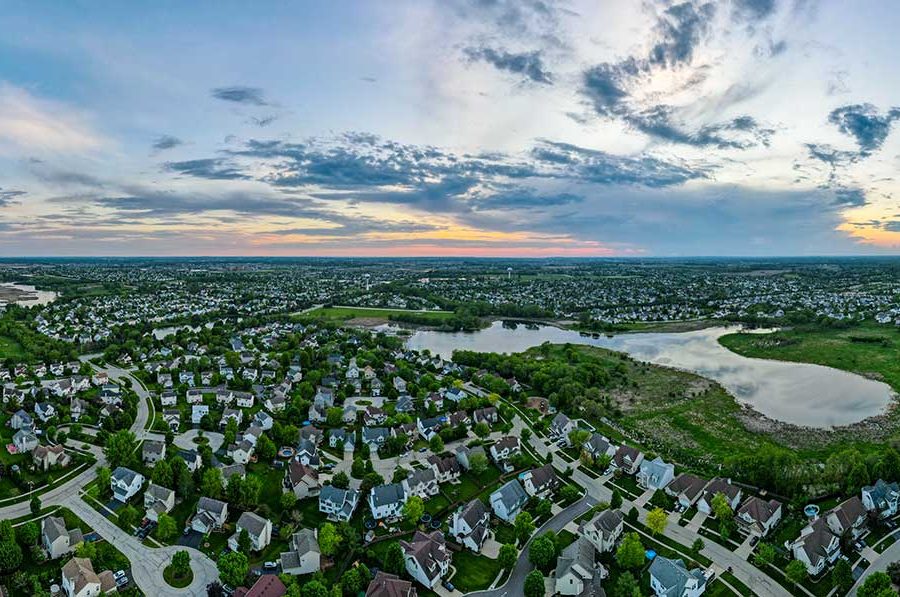 An aerial image of the city of Woodstock in McHenry County, Illinois, shows a large single-family housing development featuring several small ponds. A flat landscape of more houses extends far out into the distance.