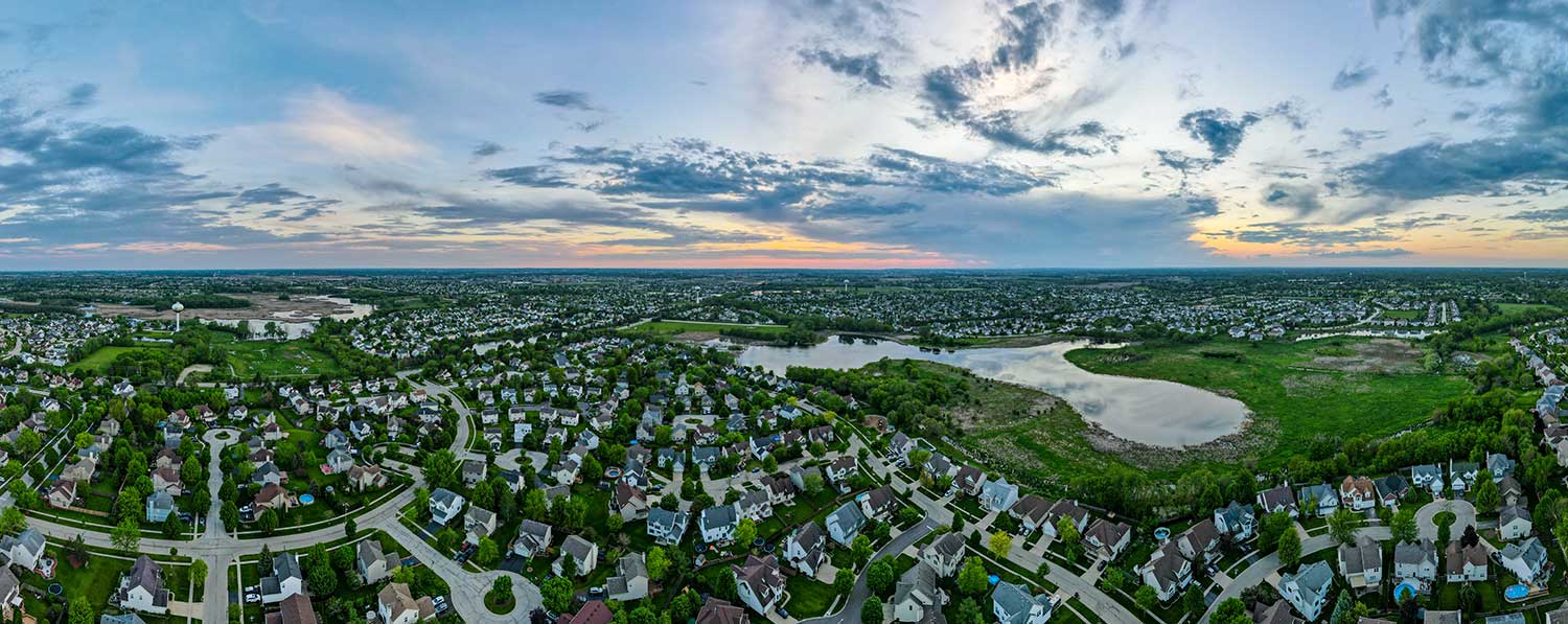 An aerial image of the city of Woodstock in McHenry County, Illinois, shows a large single-family housing development featuring several small ponds. A flat landscape of more houses extends far out into the distance.