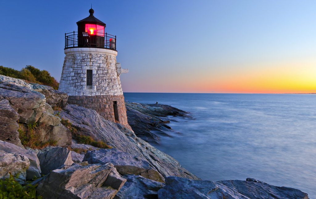 A photo of the Castle Hill Lighthouse in Rhode Island shows a squat, two-story lighthouse perched on the side of a rocky cliff overlooking the Atlantic Ocean at sunrise. The lighthouse is made of masonry stone painted light brown on the first level and white on the second level with two narrow offset windows. It features a black cupola and gallery railing, and a glowing red beacon light.
