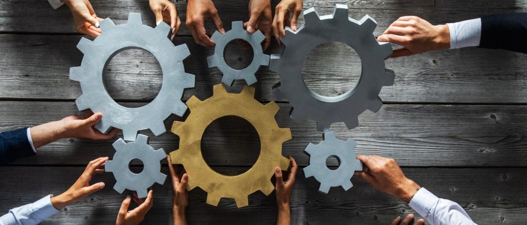 Many different people's hands hold machinery cogs of different sizes and colors together