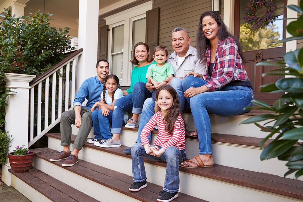 A multi-generational family including grandparents, parents, and children under age 18 sit together on the porch of their shared home.