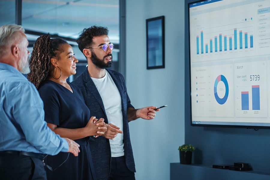 Data is presented by a consultant to two stakeholders on a large video monitor