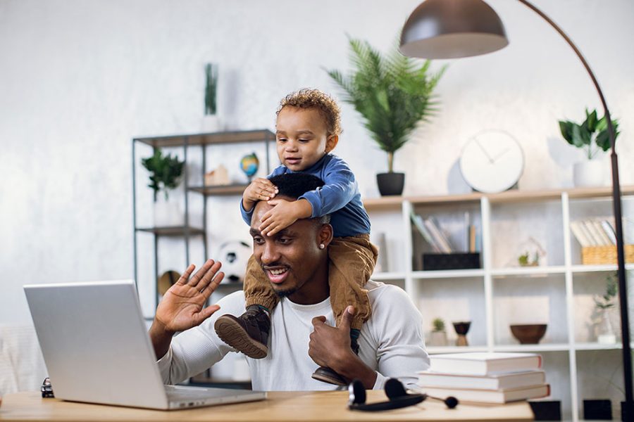 A young father with his child on his shoulders attends a virtual work meeting from home