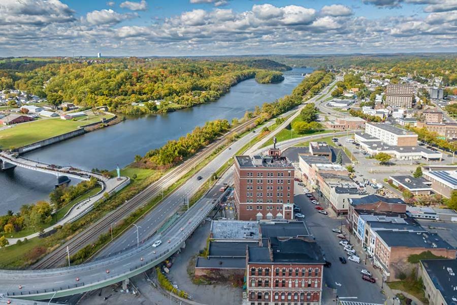 An aerial photo of the City of Amsterdam in Montgomery County shows a historic downtown area and other commercial buildings on one side of the Mohawk River, and homes, forest, and open space on the other side. An attractive bicycle/pedestrian bridge connects the two sides of the river