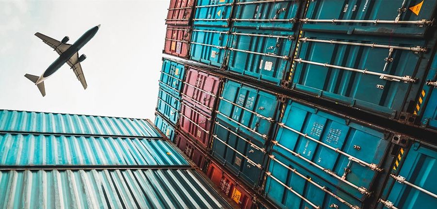 A commercial jet flies over tall stacks of larger cargo containers