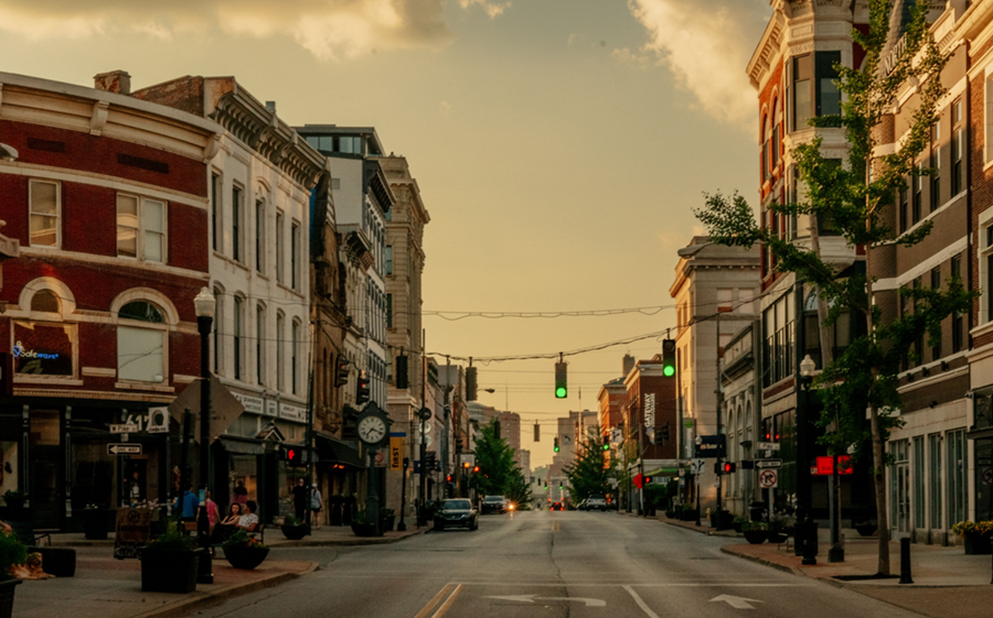 Photo of a street in downtown Covington, Kentucky, features many three- to four-story historical buildings on both sides with businesses on the first floor and offices or residential space on the upper floors. Streetscaping elements like benches, planters, bollards, trees, lampposts, and a vintage street clock are visible.