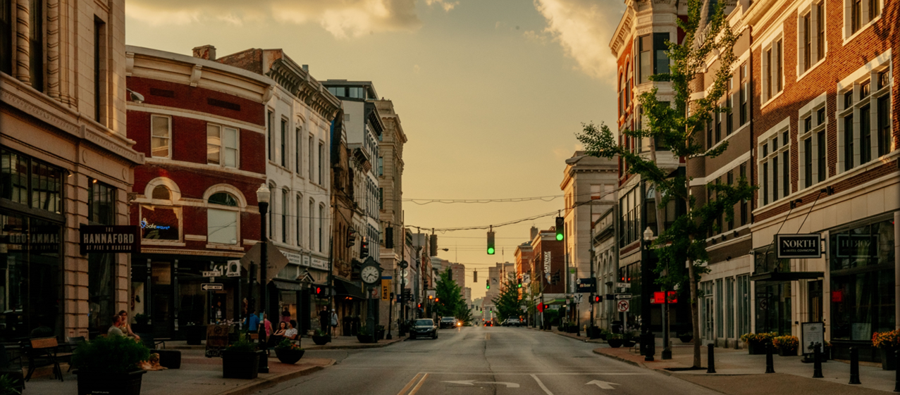 Covington KY | Camoin Associates Photo of a street in downtown Covington, Kentucky, features many three- to four-story historical buildings on both sides with businesses on the first floor and offices or residential space on the upper floors. Streetscaping elements like benches, planters, bollards, trees, lampposts, and a vintage street clock are visible. Photo is used courtesy of Sam Greenhill.