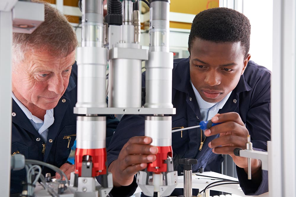 An older factory worker watches a young trainee make an adjustment to a piece of equipment on a factory assembly line