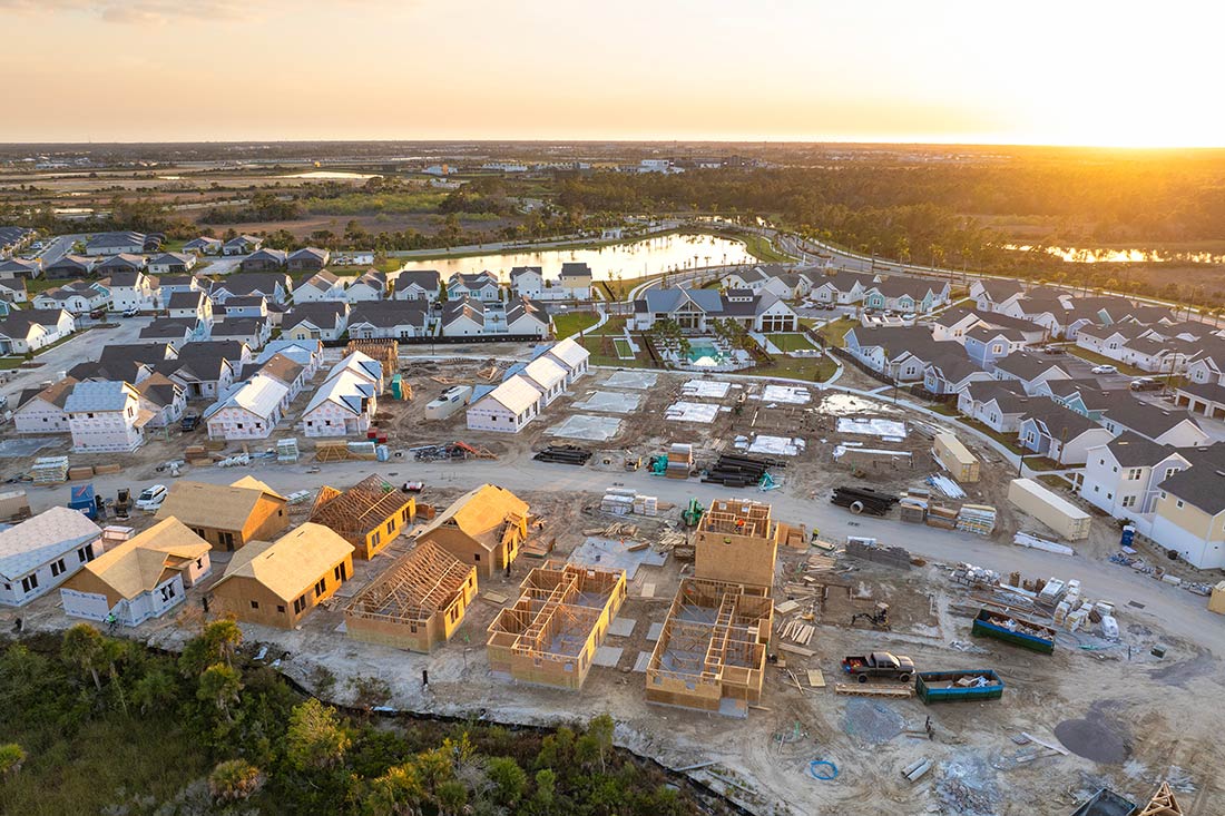 A cluster of medium-sized homes in different stages of construction on a flat piece of land surrounded by ponds and forest