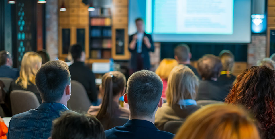 An image of a person leading a class or workshop presentation in front of a room full of participants with a large display screen behind them