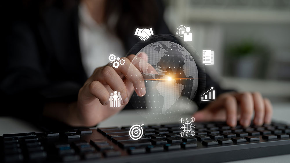 A person sits at a computer with an image of the Earth floating in front of her. She uses a digital pen to point at a spot on the Earth. Icons representing lead generation, prospecting, data, investments, and meetings surround the image of the Earth.