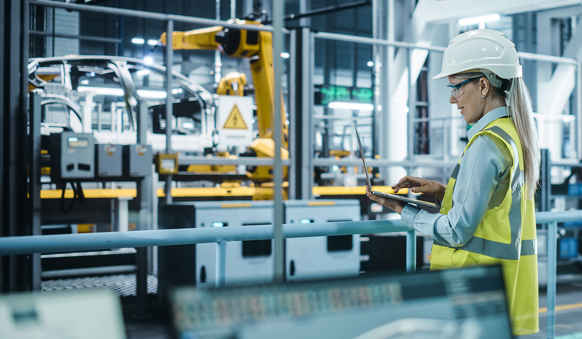 A woman wearing a yellow safety vest and a white hard hat uses a laptop computer to operate robots on an Advanced Manufacturing automobile assembly line.