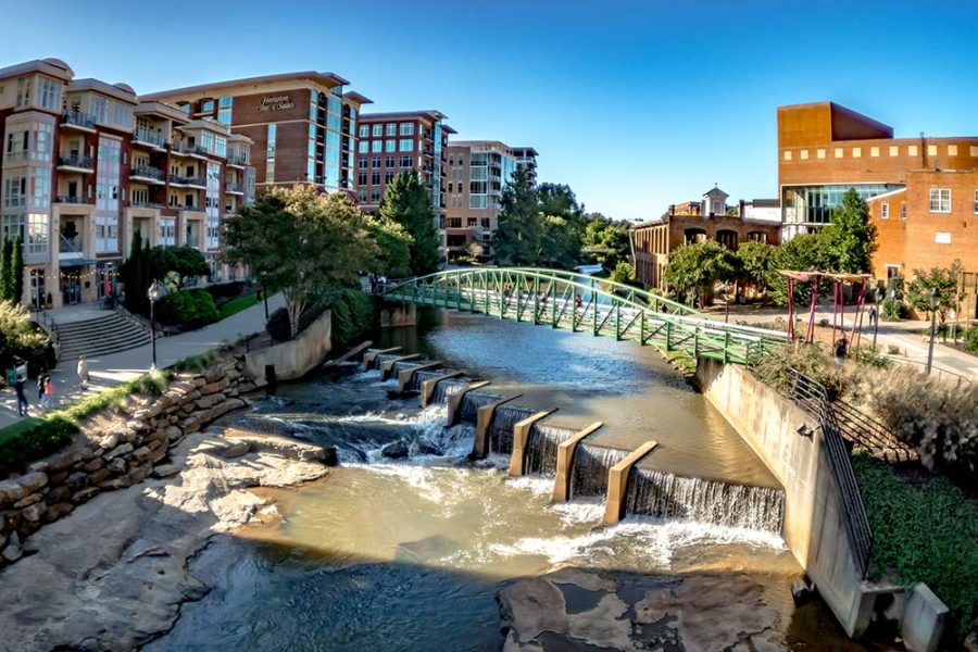 The Reedy River flows through the city of Greenville, South Carolina. Multi-story residential and commercial buildings flank both sides of the river and a green pedestrian bridge crosses it.