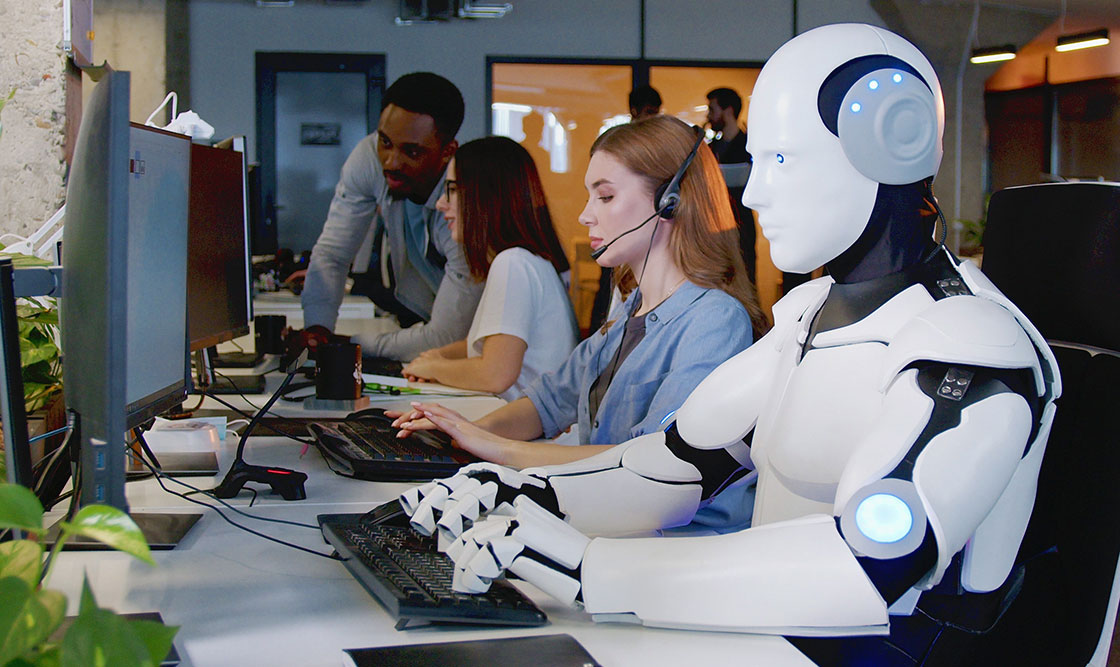 An office setting with multiple work spaces side by side shows a sleek white humanoid-looking robot working on a computer next to human co-workers
