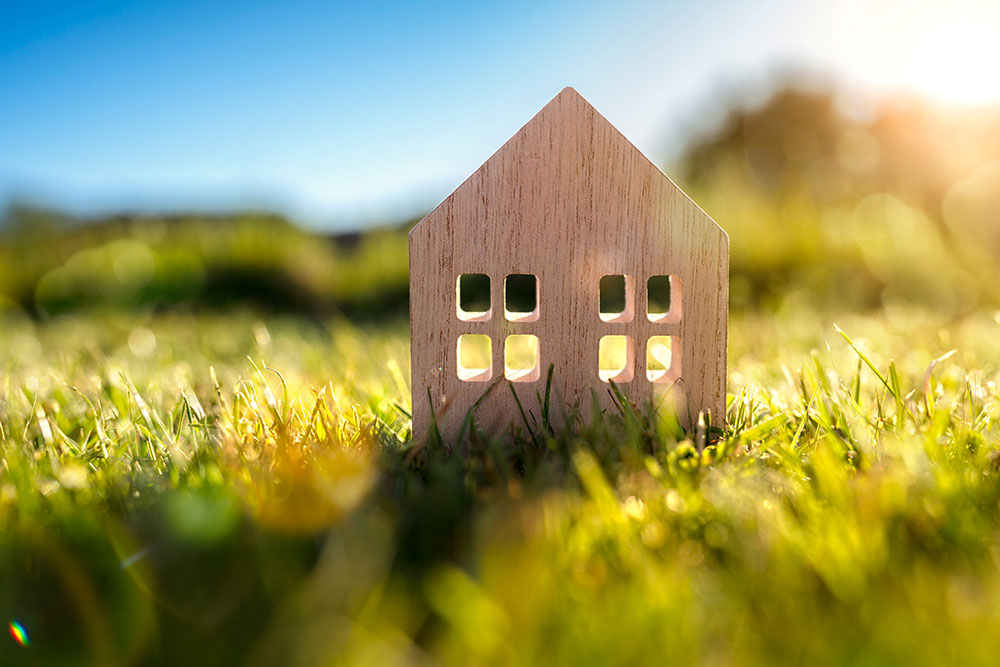 A small wooden model of a single-story house sits in a grassy field in a rural area